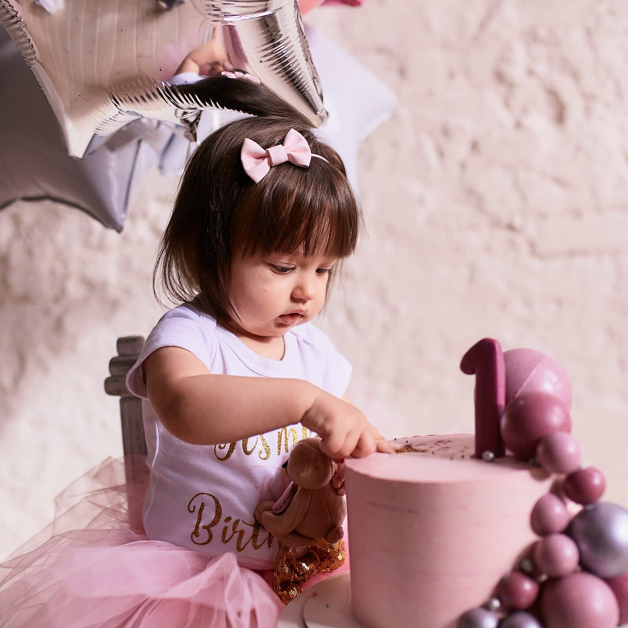 Little birthday girl. Charming baby in pink dress sits on the chair under pink, silver and white baloons and tastes delicious pink cake with cream and macaroons.