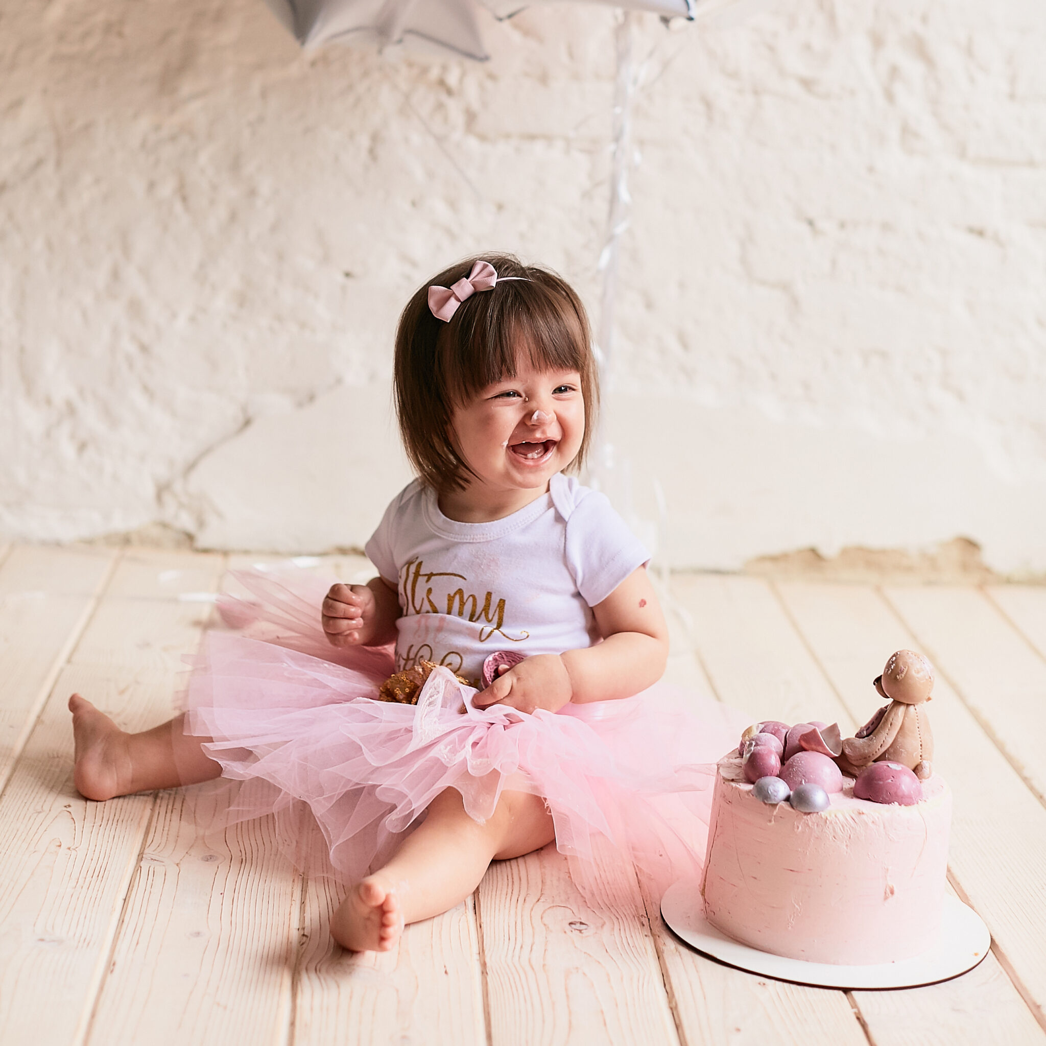 Little birthday girl. Charming baby in pink dress sits on the chair under pink, silver and white baloons and tastes delicious pink cake with cream and macaroons.