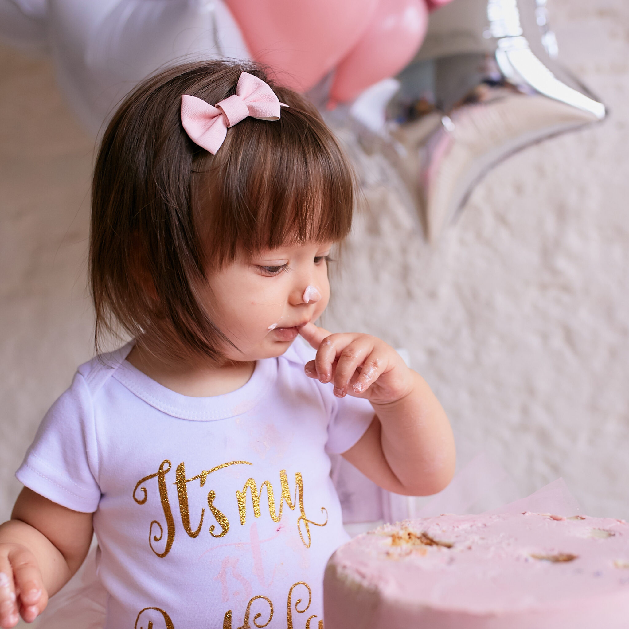 Little birthday girl. Charming baby in pink dress sits on the chair under pink, silver and white baloons and tastes delicious pink cake with cream and macaroons.