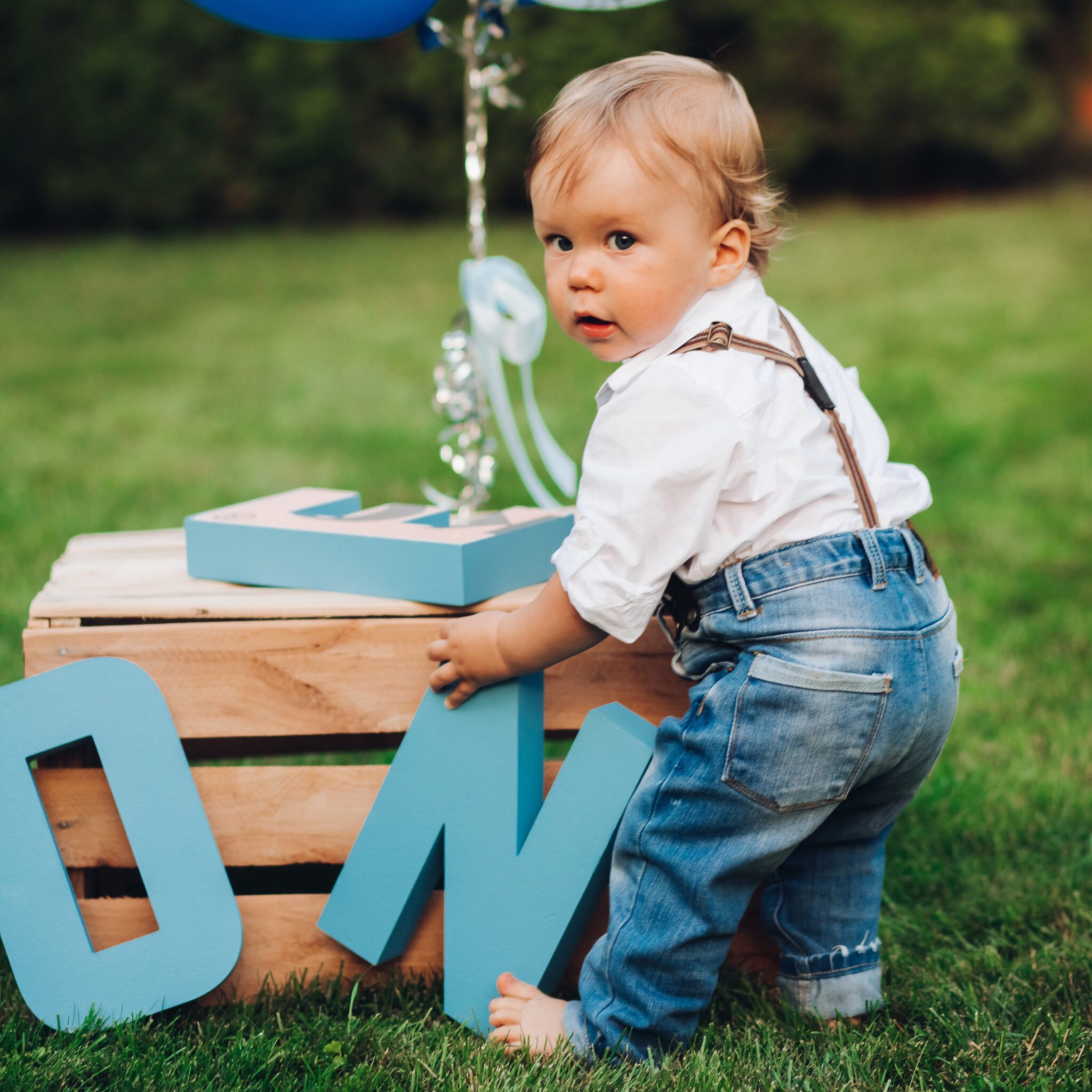 stock photo of an adorable little boy in jeans, shirt and suspenders playing with birthday decorations on the lawn in the backyard. Summer day out. Birthday concept. Air balloons and wooden letters.