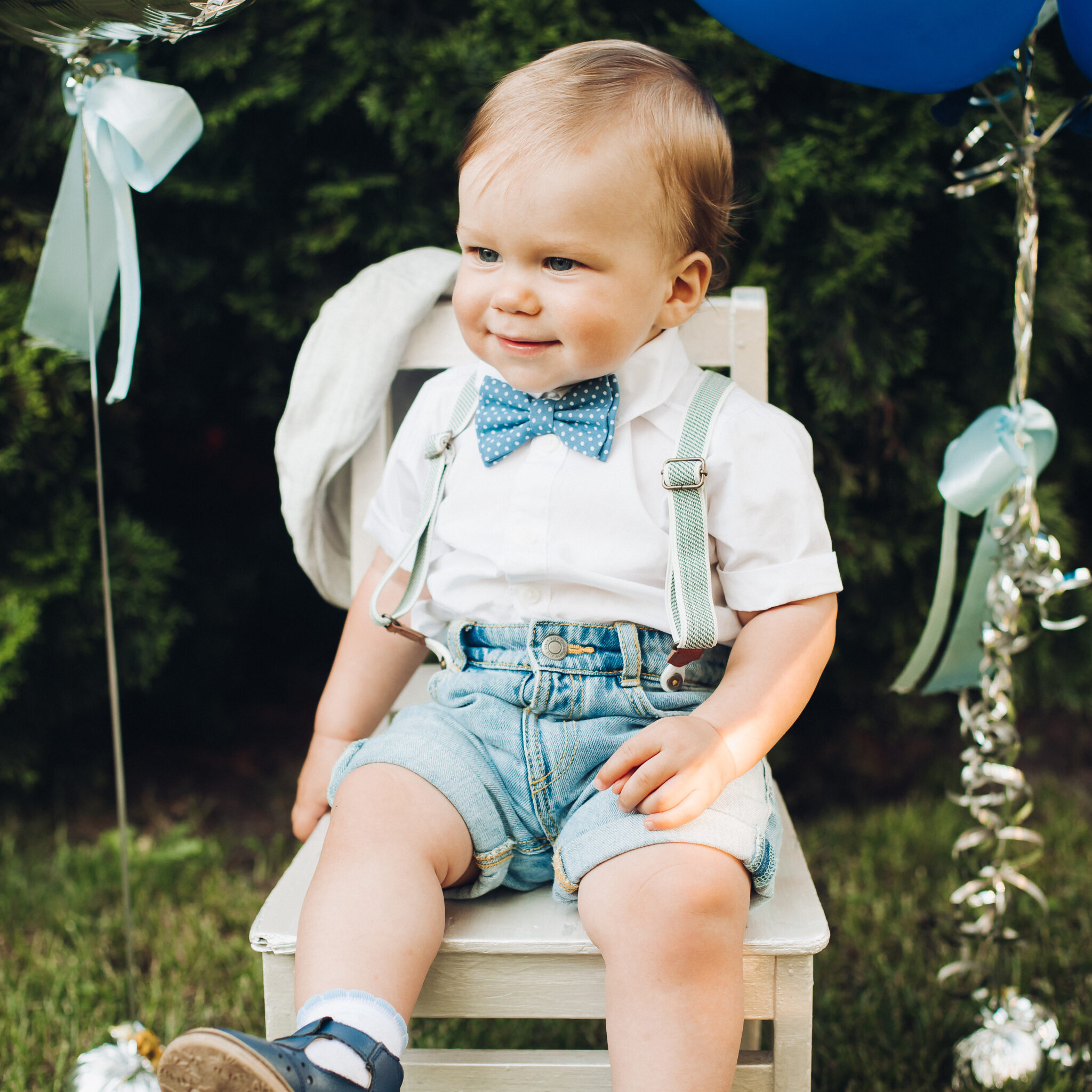 Stock photo portrait of a cute little boy wearing denim shorts, shirt, suspenders and a bow with sandals sitting on a small chair on the grass. Beautiful air balloons in the background. Child’s birthday.